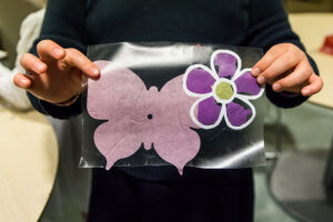 A close up a pink butterfly and purple flower blossom held up in a child's hands.