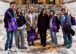A group of people pose for a photo in front of a marble staircase in a government building. Three of them hold purple tote bags with white text reading: POVERTY IS A POLICY ISSUE.