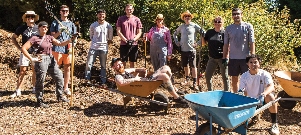 On a bright sunny day, a group of 11 people dressed in gardening clothes, some with pitchforks, pose in the midst of a large mulch pile. One person lounges in front of them in an orange wheelbarrow. Another kneels holding the handles of an empty blue wheelbarrow.