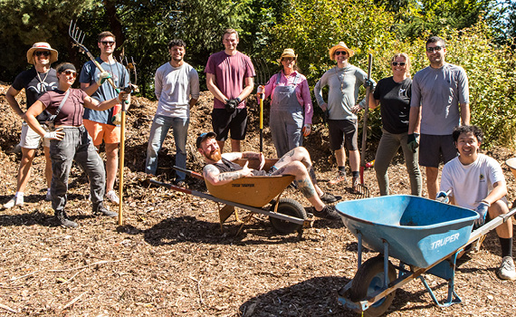 On a bright sunny day, a group of 11 people dressed in gardening clothes, some with pitchforks, pose in the midst of a large mulch pile. One person lounges in front of them in an orange wheelbarrow. Another kneels holding the handles of an empty blue wheelbarrow.