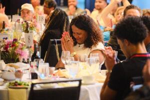 A woman at a table smiling with eyes close and clasping her hands in gratitude or prayer