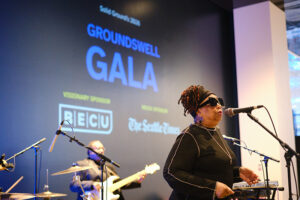 A woman singing infront of a projects that reads "Groundswell Gala"