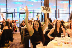 A women in a crowded ballroom holding up both arms, one of them holding a bid card