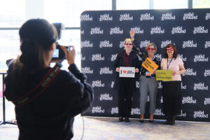 A photographer takes a picture of three people holding signs in front of a backdrop.