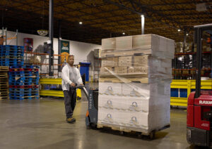A man in a grey sweatshirt uses a hand forklift to move a large pallet of packaged boxes in a massive warehouse.