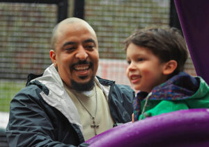 A close up of a father smiling as his young son starts down a purple slide.