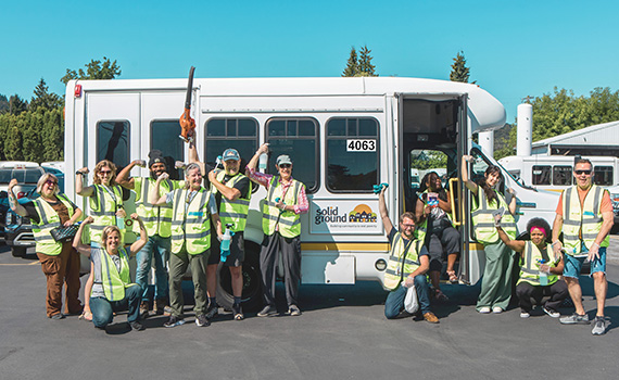 A large group of people standing in front of an accessible transportation bus, wearing neon yellow safety vests and cheering and showing their biceps as they pose for a photo.
