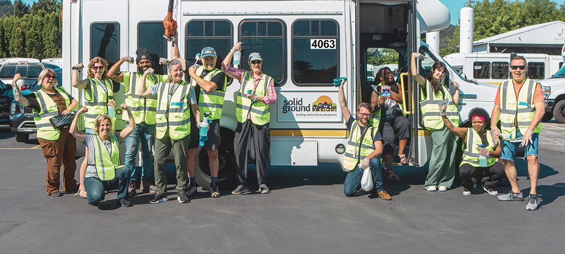 A large group of people standing in front of an accessible transportation bus, wearing neon yellow safety vests and cheering and showing their biceps as they pose for a photo.