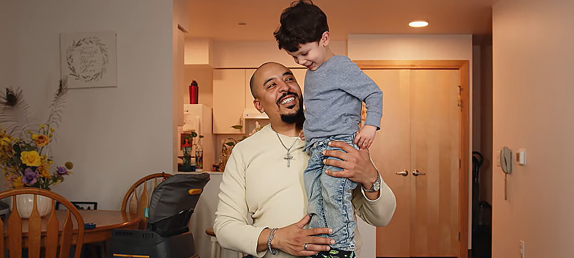 A dad sits on a chair in his apartment and beams up at his 5-year-old son, who's standing on his lap.