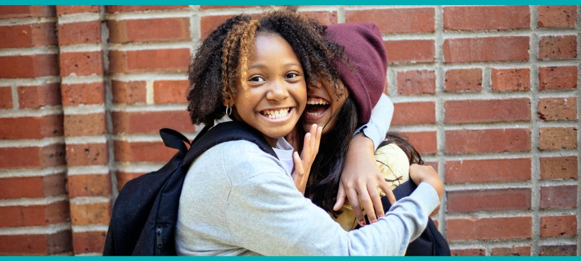 Two teen girls hug and grin. They're standing in front of a brick wall and wearing backpacks.