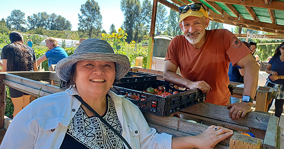 A woman in sunhat and sun-protective clothes and a man with grey facial hair, yellow cap, and rust-colored T-shirt pose by a crate of freshly harvested tomatoes at a farm stand, with a garden in the background behind them.