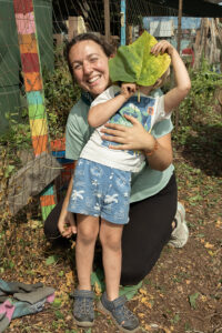 A woman holds a small child hiding their face behind a leafe
