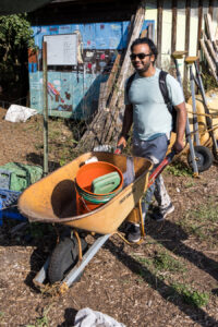 A volunteer pushing a wheelbarrow with a bucket and other gardening equipment.