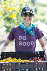 A woman wearing a shirt that reads "Do Good" holds up a crate of freshly harvested tomatoes.
