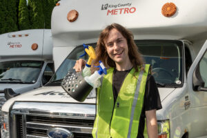 A volunteer standing in front of a paratransit vehicle holding a plastic caddy with two spray bottles.
