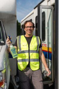 A volunteer in a reflective vest stands between two paratransit vehicles holding a spray bottle.