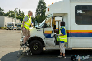 One volunteer in a reflective vest wipes down the hood of a paratransit bus while another works inside the cab.