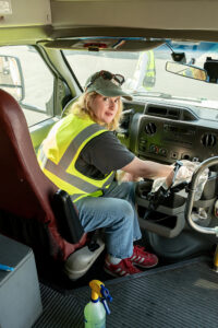 A volunteer in a reflective vest sits in the drivers seat of a paratransit bus while whiping down the dashboard with a rag.