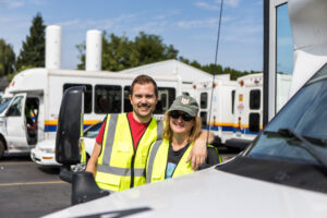 Two volunteers in reflective vests pose in front of a fleet of para transit buses.