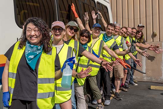 A group of smiling people wearing neon yellow safety vests line up, many with arms outstretched as if flying, next to an accessible transportation bus.