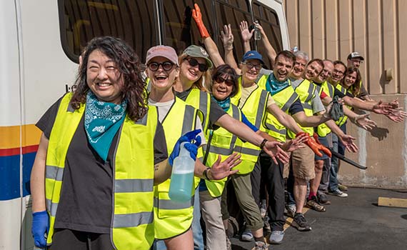 A group of smiling people wearing neon yellow safety vests line up, many with arms outstretched as if flying, next to an accessible transportation bus.