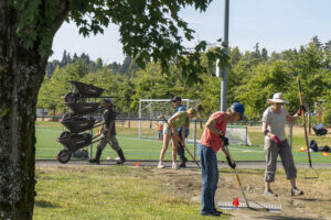Several people spread mulch while a man in the background walks by pushing five wheel barrows stacked on top of each other.