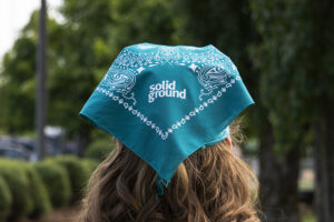 A headshot from behind of of someone wearing a teal Solid Ground bandana over their hair.