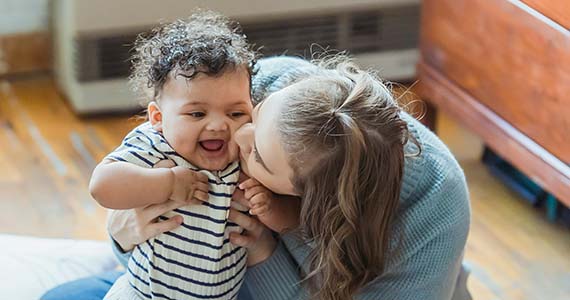 A mom in a light blue sweater and with long blondish hair kisses the cheek of her small, dark & curly-haired child, who wears a white & black striped shirt.