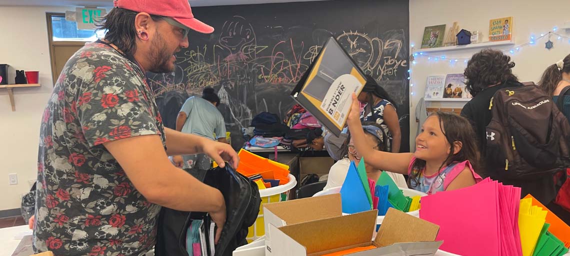 A dad in a pink cap with glasses and beard helps his daughter fill a backpack with supplies. She excitedly holds up a notebook.