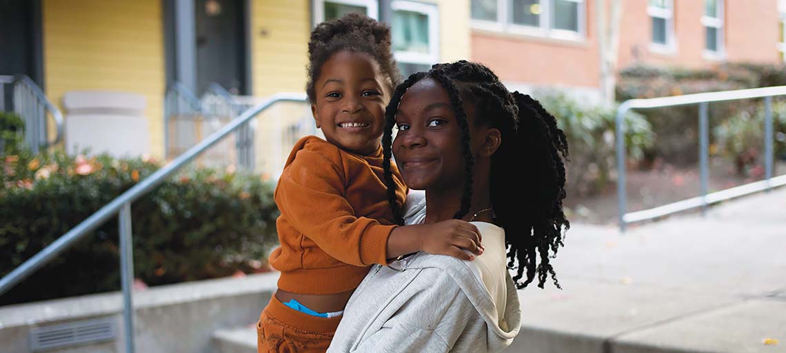 An older sister, wearing a white top, holds up her toddler sister, all dressed in orange. They smile at the camera. Behind them are yellow and peach buildings, a stairway railing, and some green bushes.